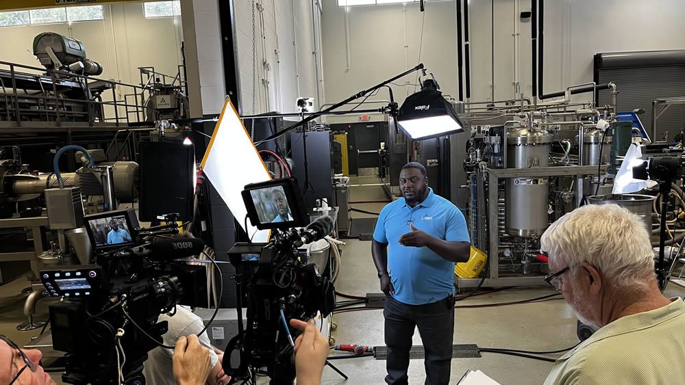 A black man in a blue shirt and black pants,standing on a studio stage being filmed by a camera
